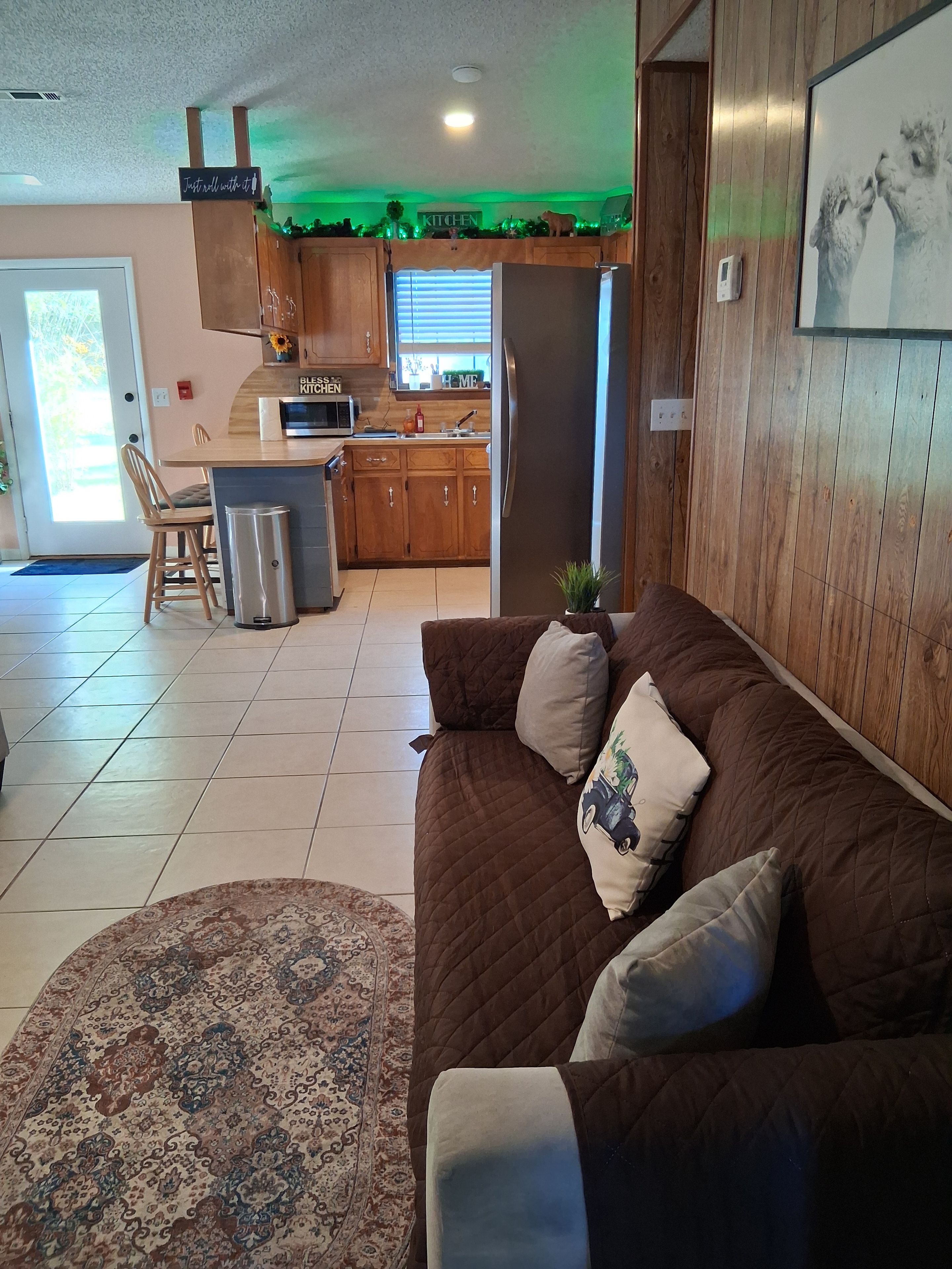 A living room with a brown sofa, a patterned rug, and a kitchen in the background.