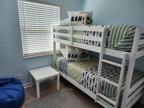 Uma cama beliche com roupa de cama estampada, uma mesinha branca pequena e um assento de saco azul em um quarto com paredes azul claro.