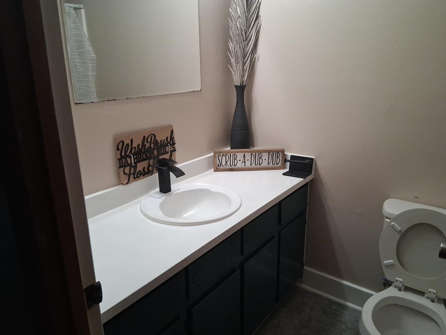 A bathroom with a white sink, a black faucet, a decorative sign, a vase with dried plants, and a toilet.