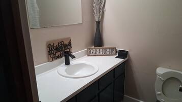 A bathroom with a white sink, a black faucet, a decorative sign, a vase with dried plants, and a toilet.