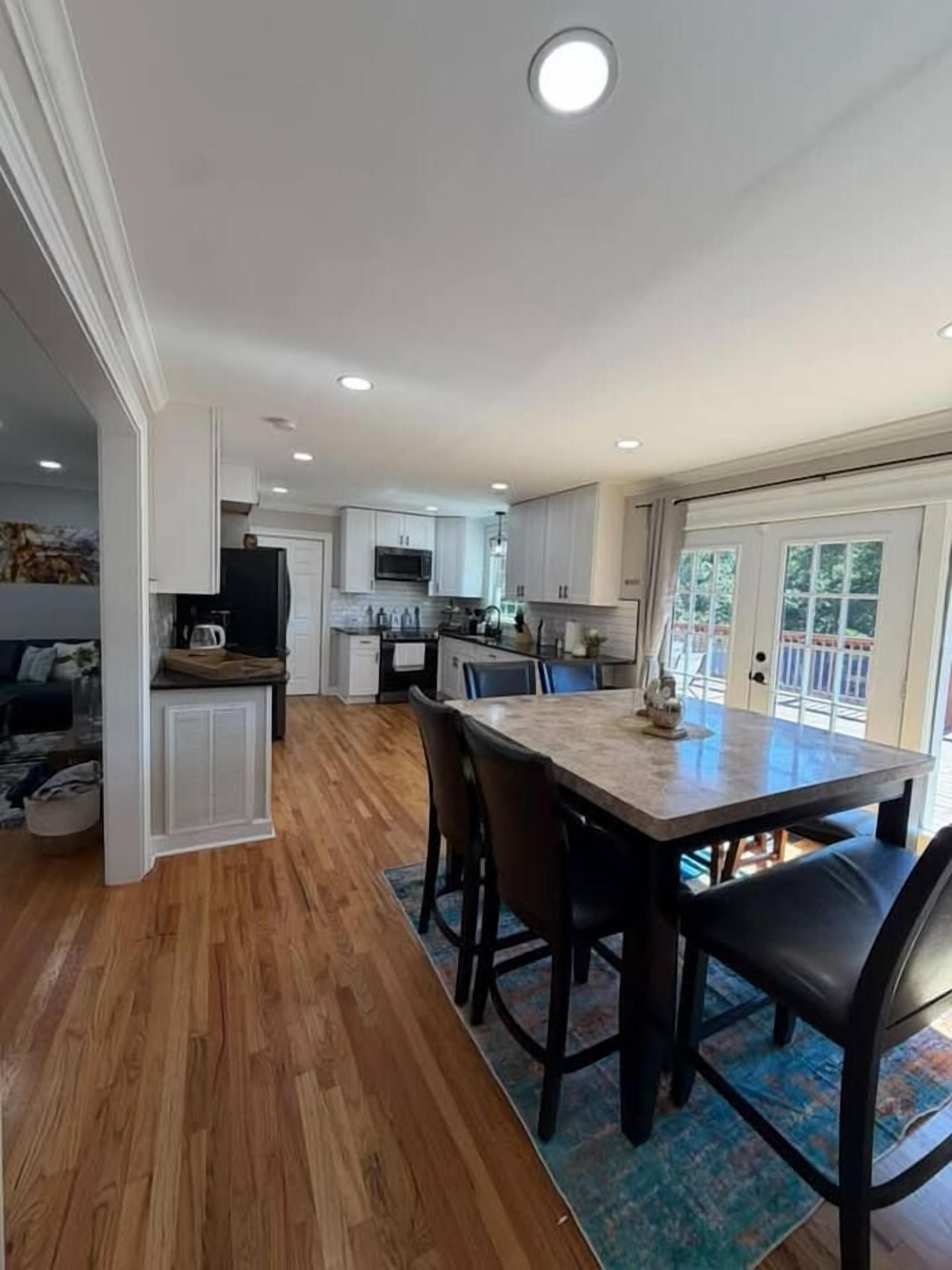 A modern kitchen with white cabinets, a dining area with a wooden table and chairs, and a view of the outdoors through a glass door.