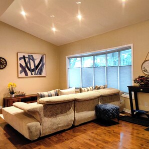 A living room with a sectional sofa, a clock, and a window with blinds. - Poolside Retreat | Private Pool, Hot Tub & Huge Deck in Niagara-on-the-Lake (Niagara-on-the-Lake)
