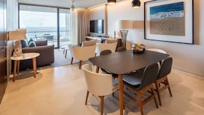 Habitación de hotel moderna con zona de comedor, un sofá, televisión y un amplio ventanal con vista al mar.