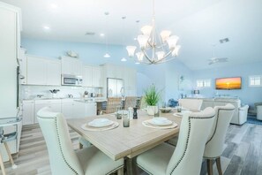 A modern kitchen with white cabinets, a dining area with a wooden table, and a living room with a TV. - RGB Pool & Game Room | Steps to Beach | 3BR Oasis (Fort Myers Beach)