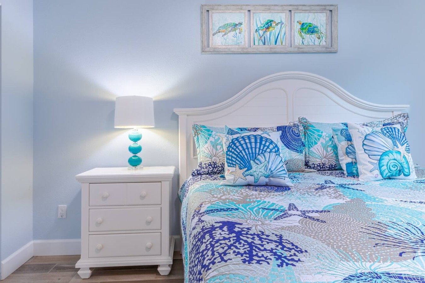 A bedroom with a white bed, a nightstand with a lamp, and a wall with three framed pictures.