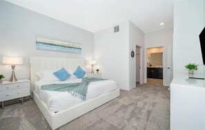 Une chambre à coucher comprenant un lit, des tables de chevet, une commode, un tableau et une porte donnant sur une salle de bain.