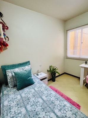 Un dormitorio con cama, mesita de noche, una planta en maceta y una ventana con persianas.