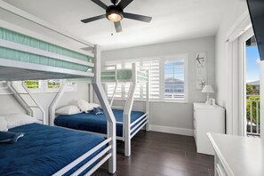 Ein Zimmer mit Etagenbetten, einem Deckenventilator, einem Fenster mit Jalousien und einem kleinen Schreibtisch mit Lampe.