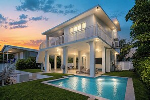 Ein zweistöckiges Haus mit überdachter Terrasse, Schwimmbecken und Blick auf den Sonnenuntergang.
