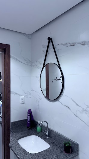 A bathroom with a marble wall, a round mirror with a black frame, a white sink, and a granite countertop.