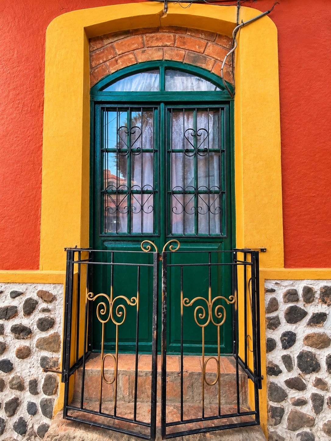 Une porte verte avec un portail en métal, encadrée dans un mur jaune et rouge dont la base est en pierre.