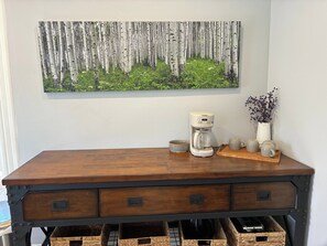 Ein Holztisch mit Kaffeemaschine, einer Blumenvase und einem Waldgemälde an der Wand.