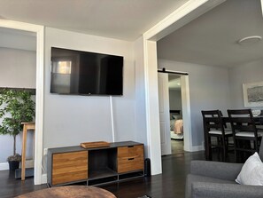 Una sala moderna con un televisor de pantalla plana montado en la pared, un mueble de entretenimiento de madera, un sofá gris y una zona de comedor con sillas de madera oscura.
