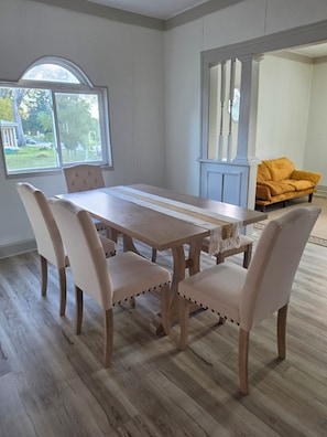 A dining room with a wooden table and chairs, a window with a view of the outdoors, and a yellow sofa in the background. - Cozy 2-Bedroom Home | Near Riverside Park (Indianapolis)