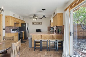 A compact kitchen with wooden cabinets, a central island with bar stools, a ceiling fan, and a view of a tree through a glass door. - Tranquil Lake Havasu Getaway - Private Pool - Rooftop Patio (Lake Havasu City)