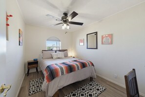 Une chambre à coucher comprenant un lit, un ventilateur de plafond, une table de chevet, une horloge, une fenêtre et des œuvres d’art accrochées aux murs.