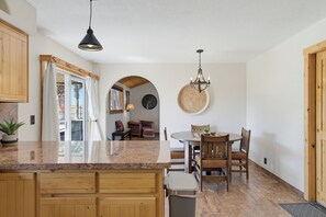 Une cuisine avec des meubles en bois, un plan de travail en granit, une table à manger avec des chaises, un lustre et une grande horloge murale.