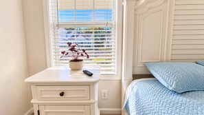 A bedroom with a bed, a nightstand, a window with blinds, and a potted plant. - The Happy Habitat (Clarksville)