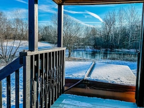 Een houten terras met een metalen hekwerk met uitzicht op een besneeerd landschap, inclusief water en bomen.