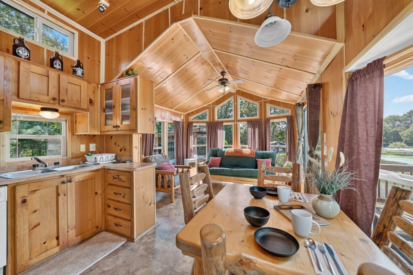 A wooden cabin interior with a kitchen, dining area, and a cozy seating area with a view of the outdoors.
