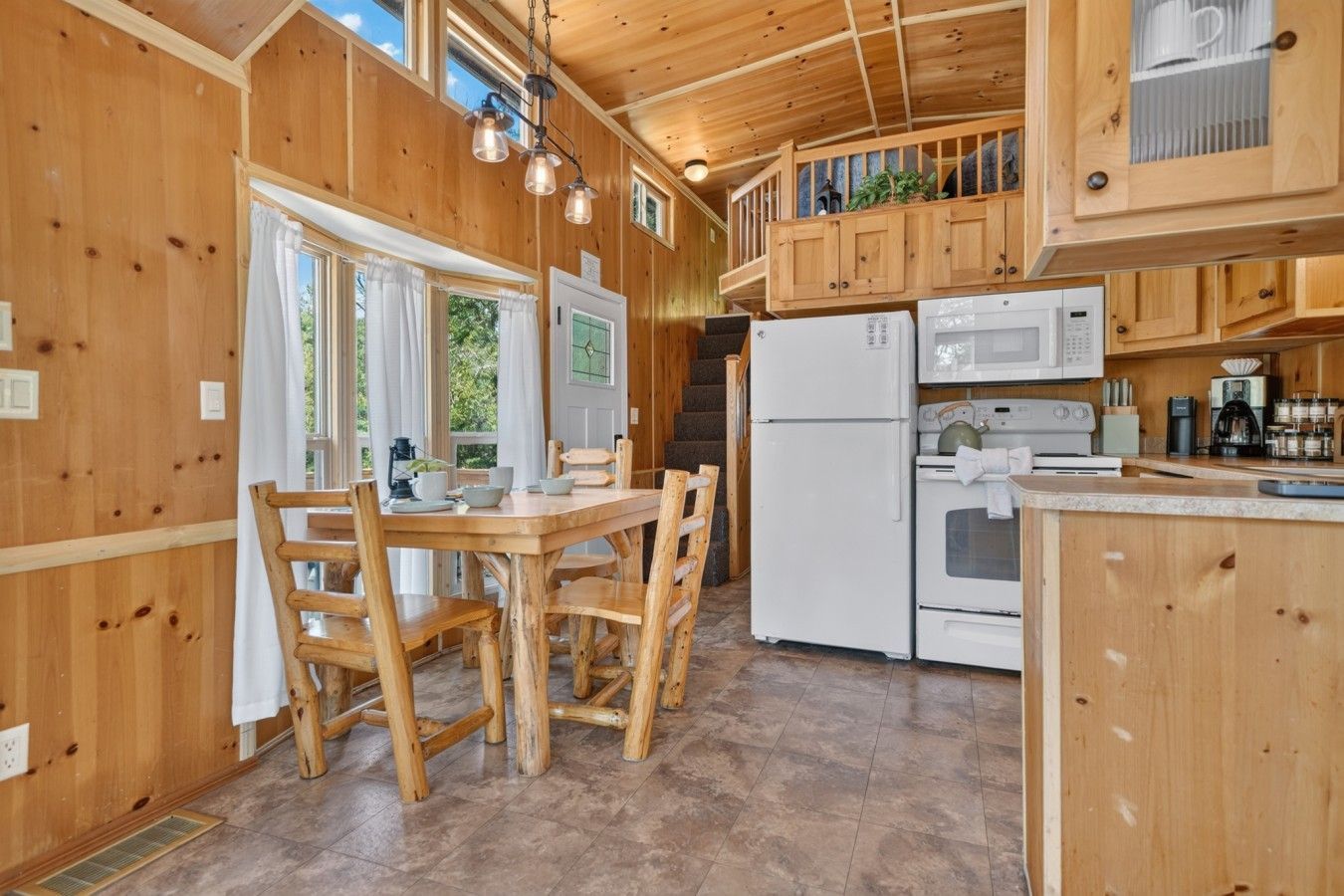 A wooden cabin interior with a dining area, kitchen, and a staircase leading to an upper level.