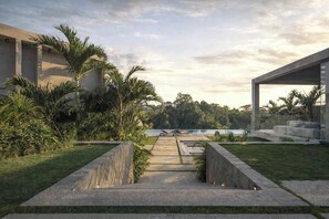 A pathway leading to a pool with a view of a lake and trees, surrounded by greenery and modern architecture. - Predmet KabaKaba Resort (Tabanan)