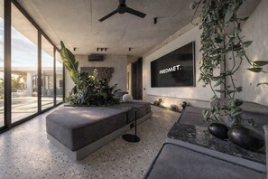 A modern living room with a large window, a grey sofa, a coffee table, and a wall-mounted TV displaying 'PREDMET I.' - Predmet KabaKaba Resort (Tabanan)