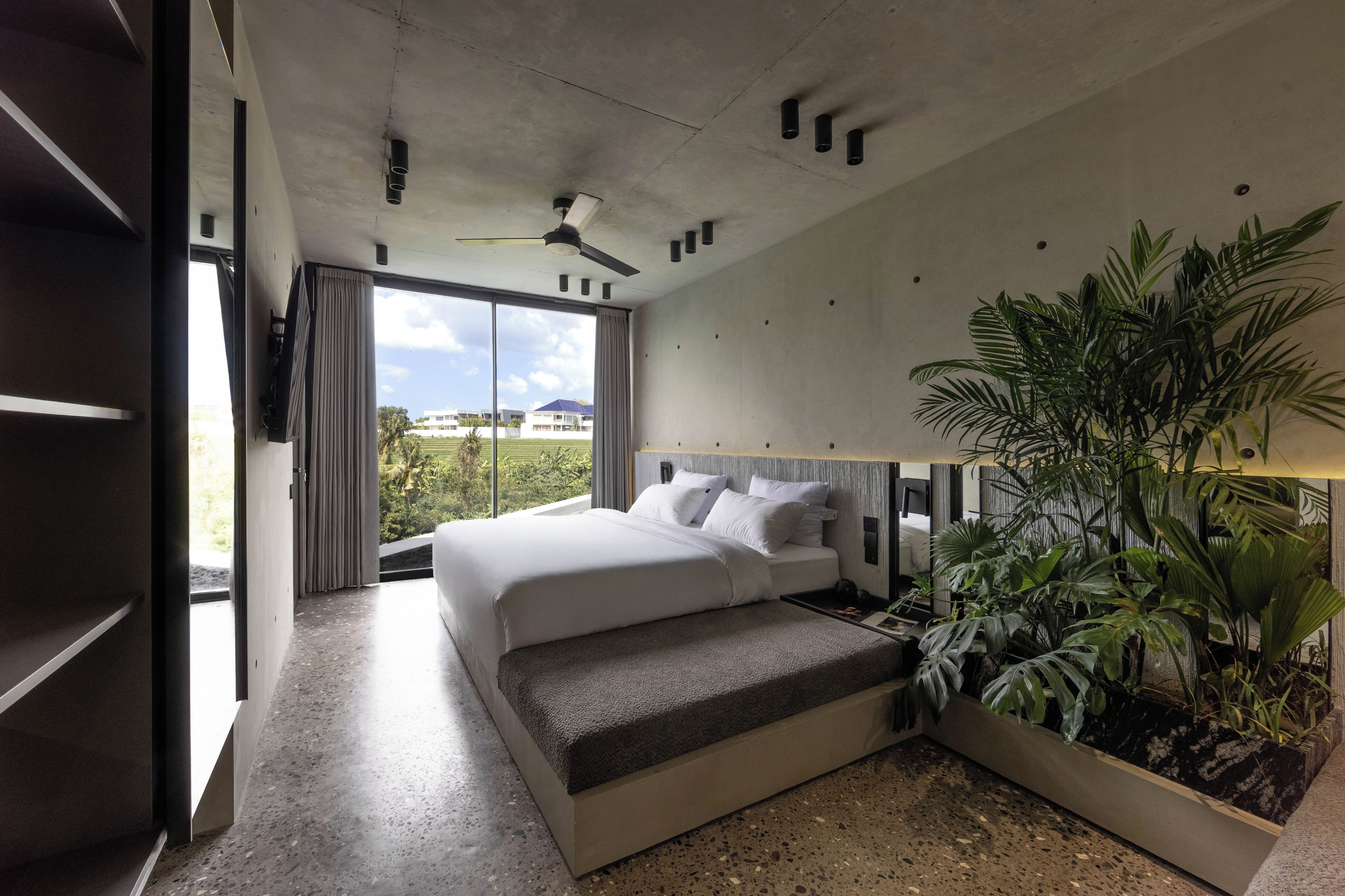 Quarto de hotel moderno com cama grande, ventilador de teto, vista para o exterior e planta em vaso.