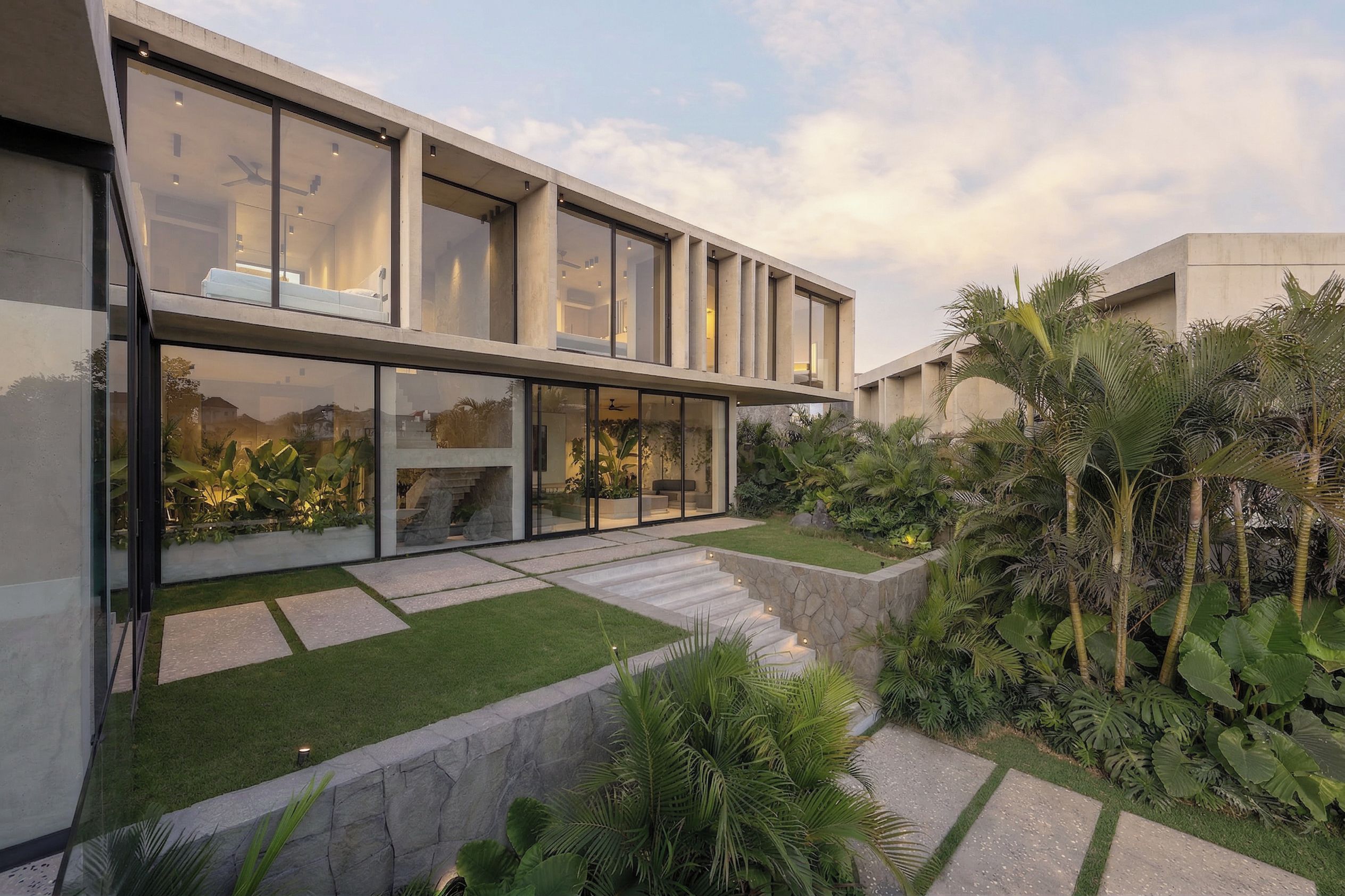 A modern two-story house with large glass windows, a stone pathway, and a well-maintained garden with tropical plants.
