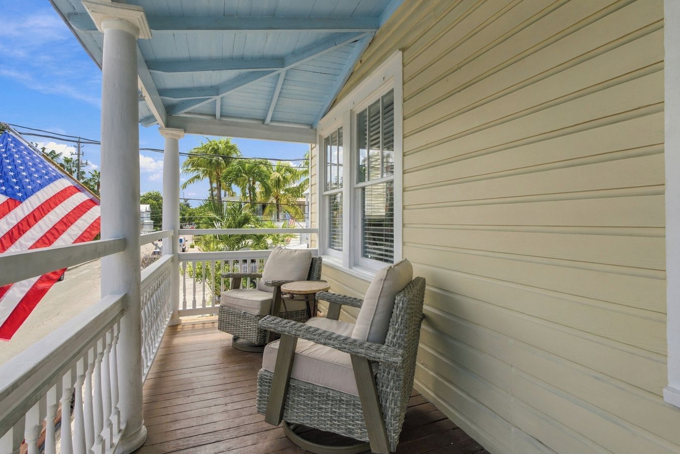 A covered porch with wicker furniture, an American flag, and a view of palm trees.