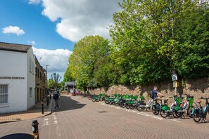 A street with a row of parked bicycles, a white building with a sign, and a tree-lined wall. - Elegant 2-bed Duplex 2 En-suites Belsize Park (London)