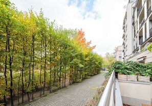 Un chemin pavé, bordé d’arbres aux feuilles vertes et jaunes, mène à un bâtiment doté de balcons.