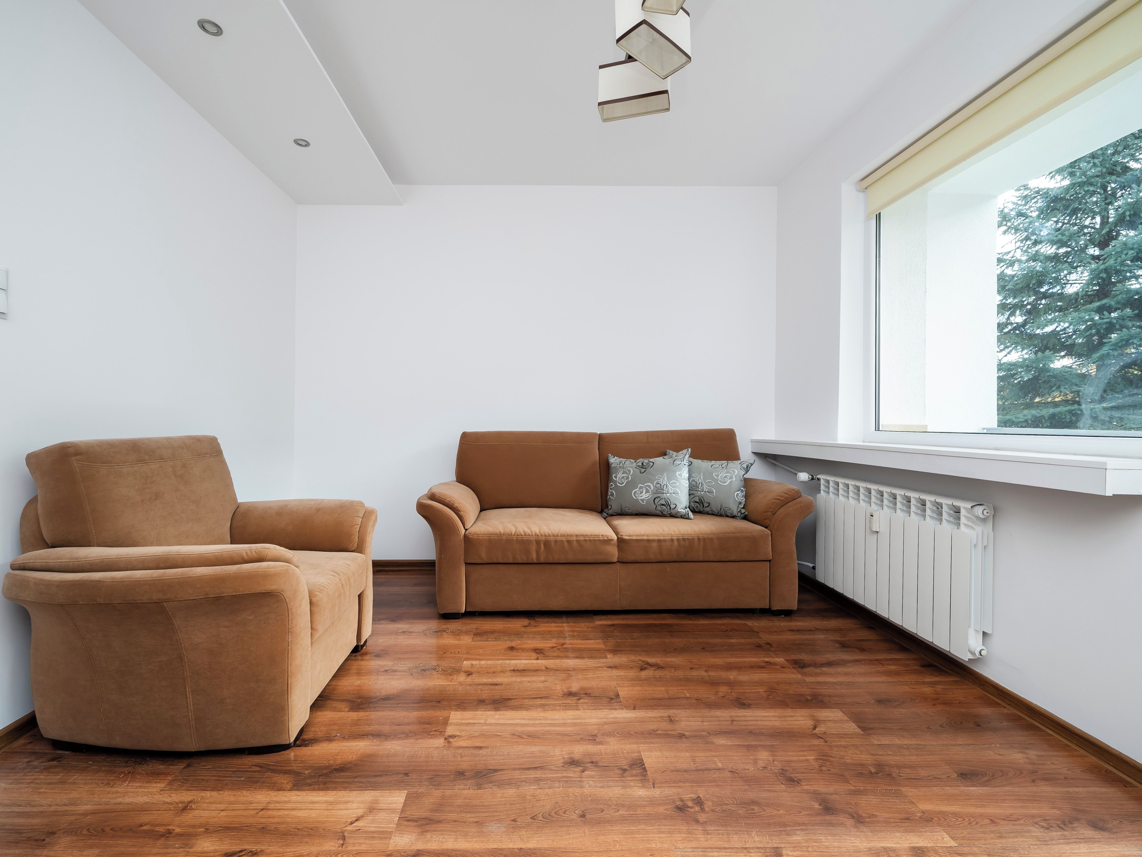 Un salon moderne avec un canapé et un fauteuil marron, un radiateur et une fenêtre donnant sur des arbres.