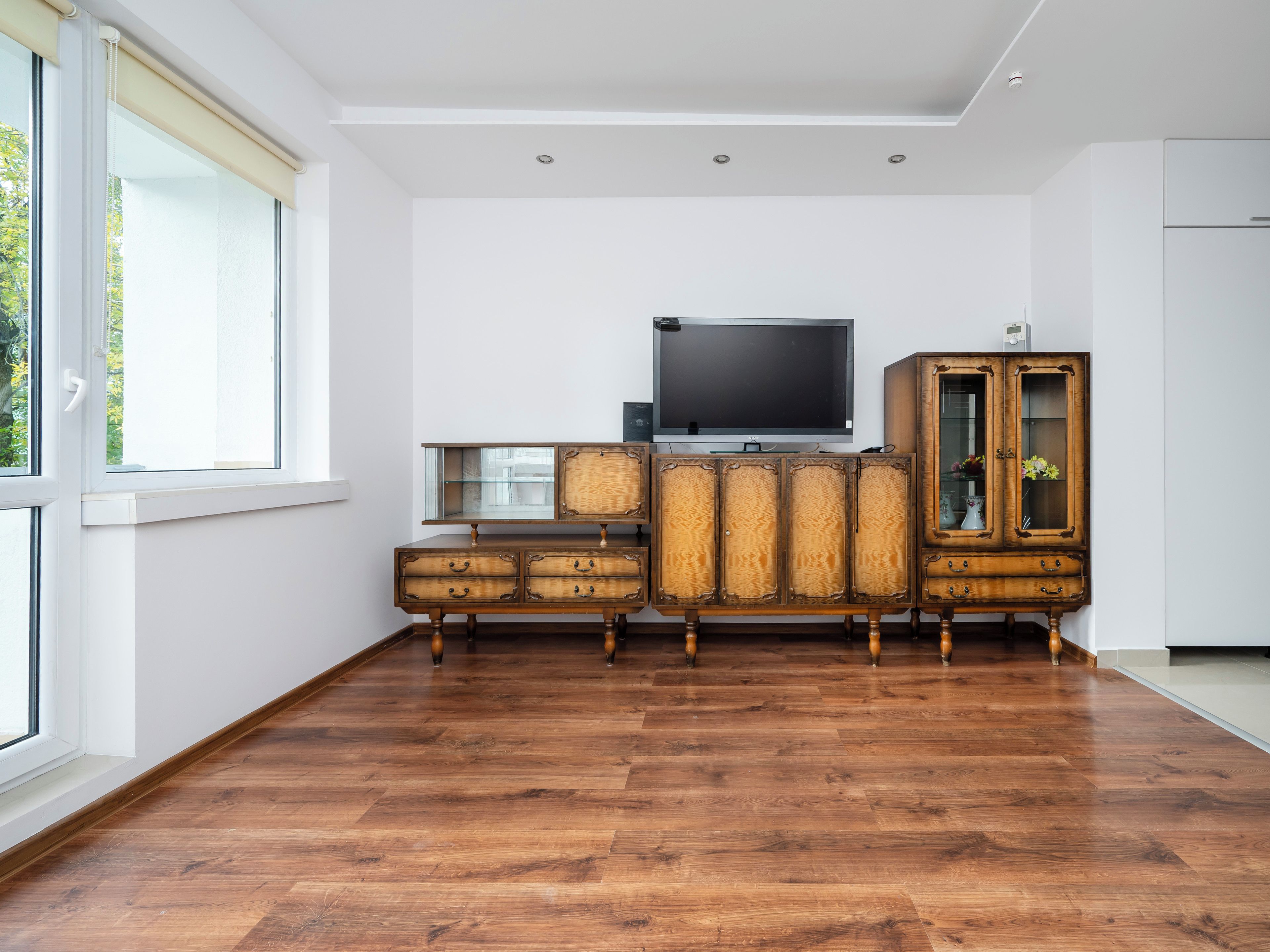 Una habitación con un ventanal grande, un aparador de madera con cajones, un televisor y un mueble con puertas de cristal.