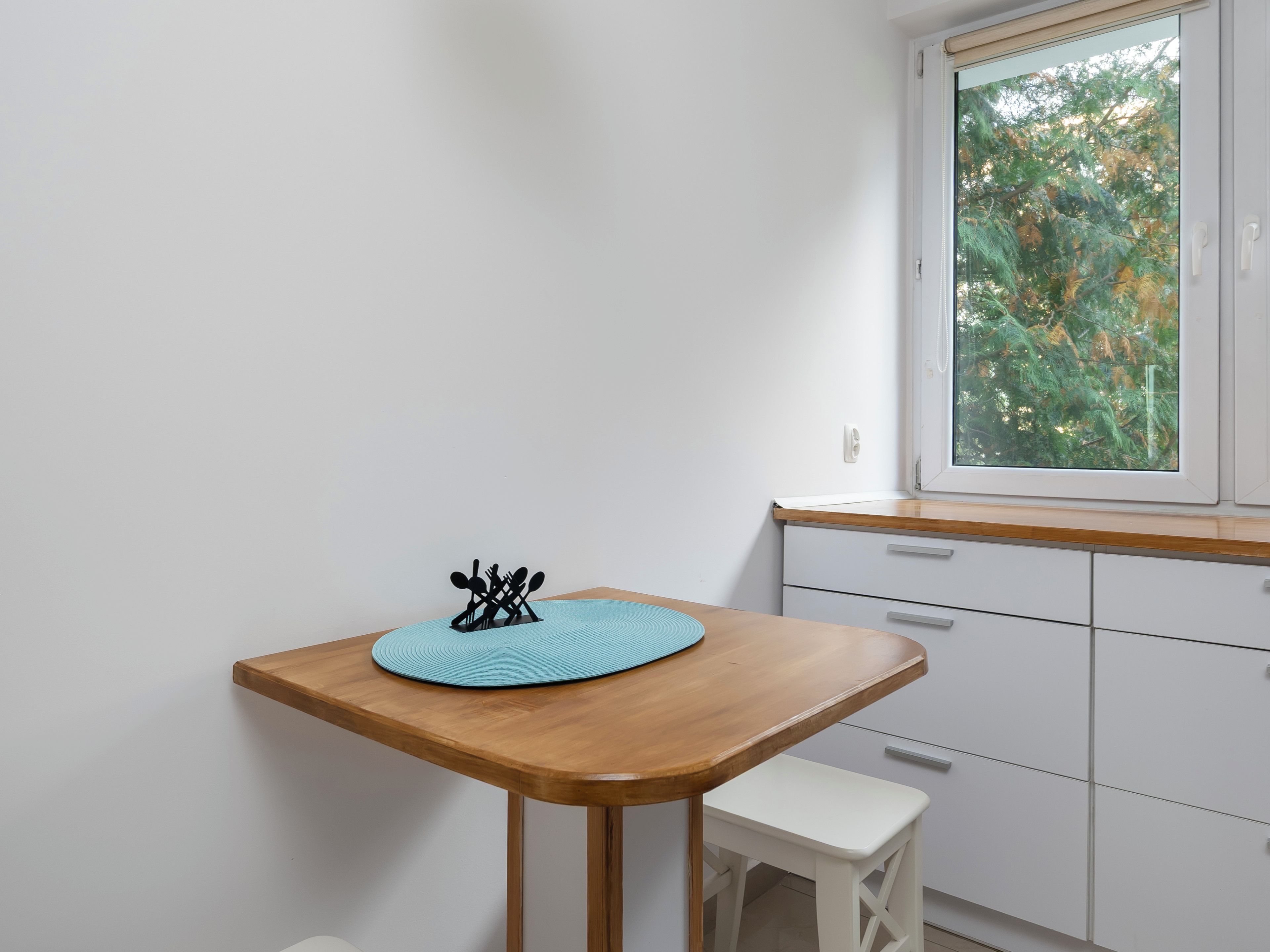 Una cocina pequeña con una mesa de madera, un tapete azul y una ventana con vistas a árboles.