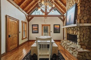 A dining area with a wooden table and chairs, a stone fireplace, and framed artwork on the walls. - Dual Blue Ridge Cabins, sleeps 14, large yard, dogs welcome! (Blue Ridge)