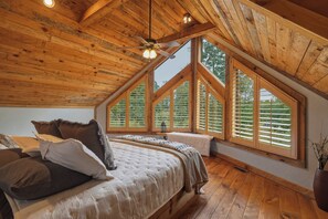 A bedroom with a sloped ceiling, wooden beams, a bed with pillows, a bedside table, and large windows with blinds. - Dual Blue Ridge Cabins, sleeps 14, large yard, dogs welcome! (Blue Ridge)