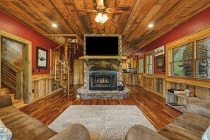 A cozy living room with a stone fireplace, wooden floors, a ceiling fan, and a staircase. - Dual Blue Ridge Cabins, sleeps 14, large yard, dogs welcome! (Blue Ridge)
