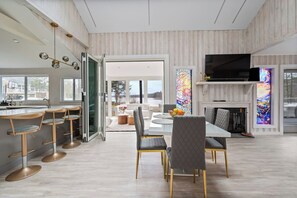 A modern kitchen with a dining area, a TV mounted above the fireplace, and a view through the glass doors. - Lakeshore Retreat-Modern Lakefront Home w/ Hot Tub (Monticello)