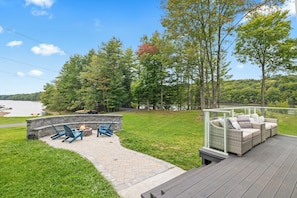 A deck with outdoor seating and a fire pit, overlooking a lake and forested area. - Lakeshore Retreat-Modern Lakefront Home w/ Hot Tub (Monticello)