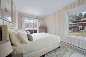 A bedroom with a large bed, a dresser, a window with curtains, and a view of the outside. - Lakeshore Retreat-Modern Lakefront Home w/ Hot Tub (Monticello)