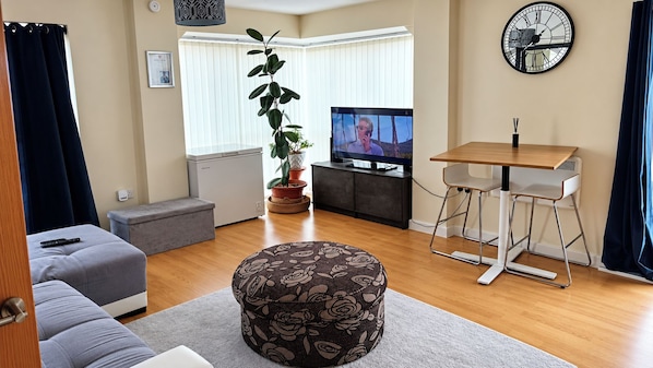 Un salon comprenant un canapé, un pouf, un téléviseur, une table à manger et des plantes en pot.