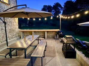 Terrassa exterior amb taules i bancs de fusta, llums penjants i una barbacoa.