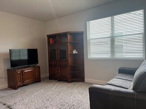 A living room with a flat-screen TV, a wooden cabinet, a gray sofa, and a window with blinds. - LUX Mins to HCA, Advent & Gemini Springs Beautiful 4 bedroom brand new home (DeBary)
