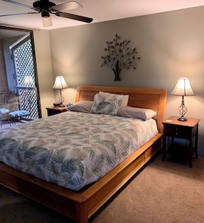 A bedroom with a bed, two bedside tables with lamps, a ceiling fan, a decorative wall piece, and a view through a window with a lattice. - Wailea Ekahi 21D top floor ocean view 1bedroom 2bath short level walk to beach! (Kihei)