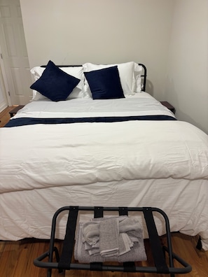 A neatly made bed with white and navy bedding, a small bedside table, and a metal drying rack with towels. - Private bathroom + bedroom UNC & Duke Hospital (Durham)