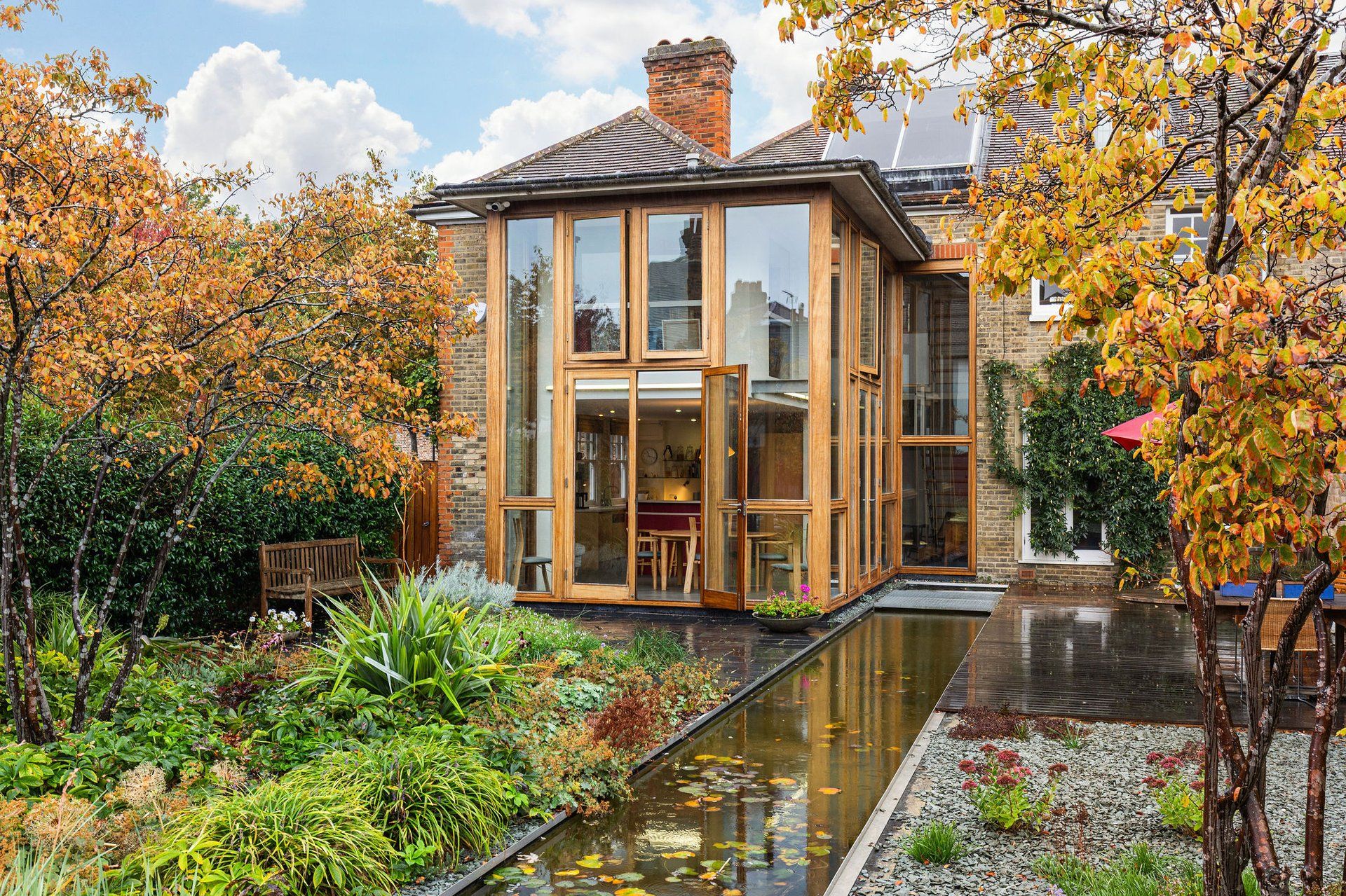 A modern house with large glass windows, a garden with a pond, and a brick chimney.