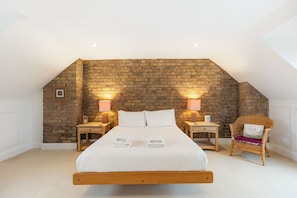 Ein Schlafzimmer mit schrägverdachter Decke, einer Ziegelwand, einem hölzernen Bett, zwei Nachttischen mit Lampen und einem Korbstuhl.