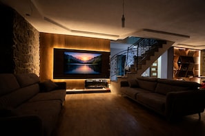 Ein modernes Wohnzimmer mit einem großen Flachbildfernseher an einer Holzwand, zwei Sofas, die dem Fernseher zugewandt sind, und einer Treppe mit Metallgeländer, die zu einer oberen Ebene führt.