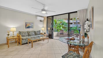 A living room with a sofa, coffee table, and chairs, a balcony with a glass door, and a ceiling fan.